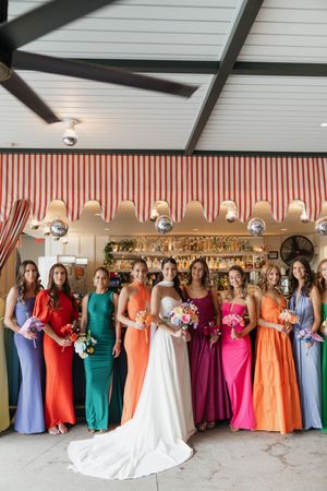 Bride in white gown poses with colorful bridesmaids in rainbow dresses holding bouquets at wedding venue.