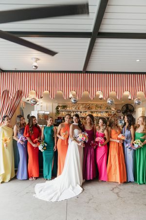 Bride in white gown poses with colorful bridesmaids in rainbow dresses holding bouquets inside a striped venue.