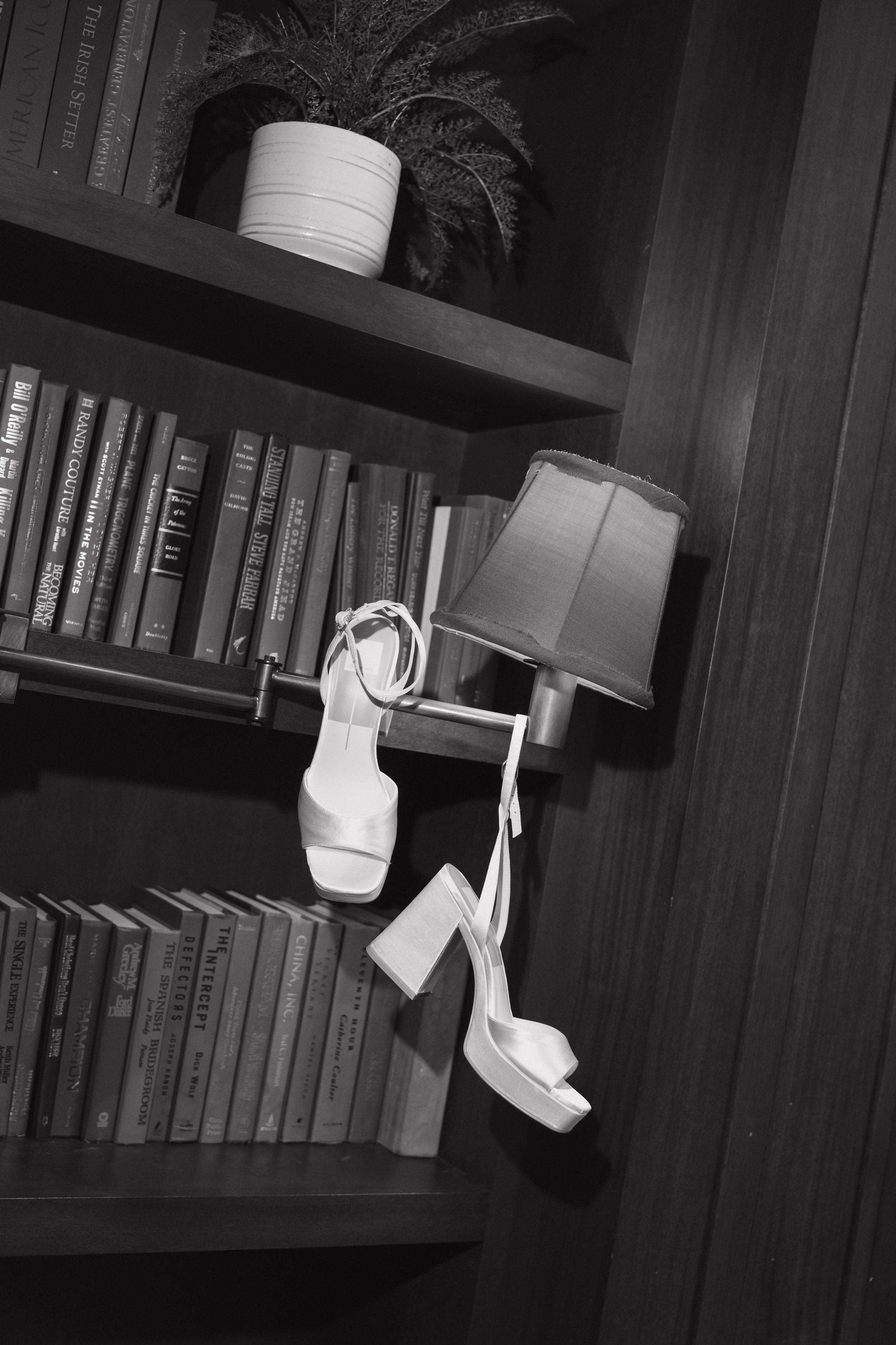 White bridal heels hanging on a lamp beside a wooden bookshelf filled with books and a potted plant.