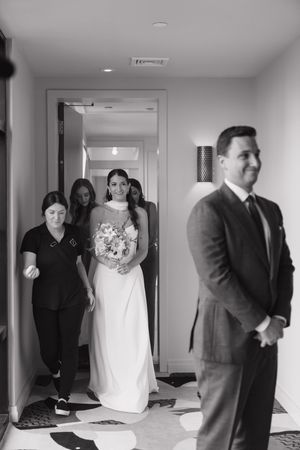 Black and white photo of a bride holding a bouquet walking down a hotel hallway toward a groom during a wedding first look.