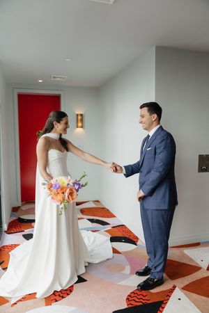 Bride in white gown holds groom's hand during first look on colorful patterned hotel floor.