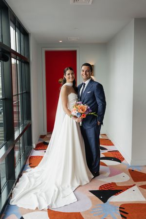 Bride and groom posing together in modern hotel hallway with colorful patterned floor and red door.