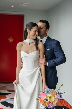 Bride in strapless white gown holds colorful bouquet as groom in navy suit kisses her cheek tenderly.