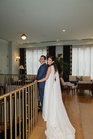 Bride in white gown with cape and groom in navy suit posing on hotel staircase balcony.