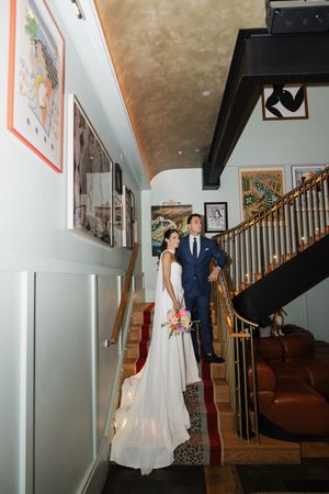 Bride and groom posing on staircase inside elegant hotel with eclectic art-lined walls.