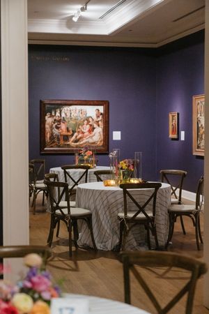 Elegant museum gallery dining setup with round tables, cross-back chairs, floral centerpieces, and classic paintings on purple walls.