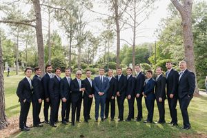 Groom with large groomsmen party in navy suits posing outdoors among tall pine trees.