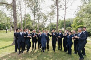 Groom and groomsmen in navy suits celebrating outdoors among tall pine trees at a wedding.