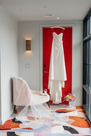 White wedding dress hanging on red door with veil draped over chair, bridal shoes and floral bouquet nearby.
