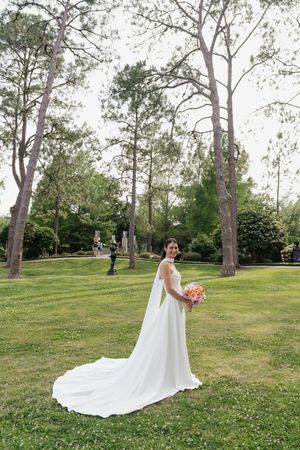 Bride in white gown with long train holds colorful bouquet in a lush green park setting.