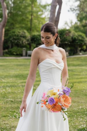 Smiling bride in a white halter gown holding a colorful bouquet of orange and purple flowers in a lush green garden.