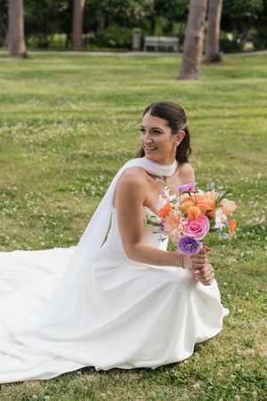 Smiling bride in white halter gown sits on green grass holding colorful orange and purple wedding bouquet outdoors.