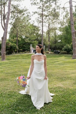 Bride in a strapless white halter gown holding a colorful bouquet while standing on green grass in a park.