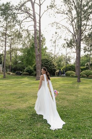 Bride in elegant white halter gown with flowing train walks through lush green park holding colorful bouquet.