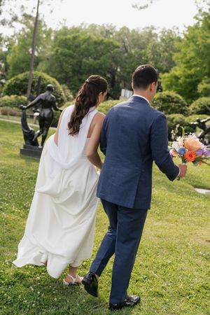 Bride and groom walk hand-in-hand through lush garden with bronze sculptures, groom carrying vibrant bouquet.