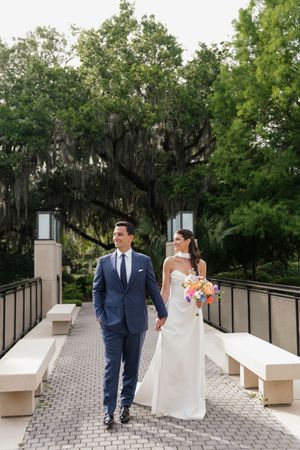 Bride and groom walk hand-in-hand on bridge surrounded by moss-draped trees, bride holding colorful bouquet.