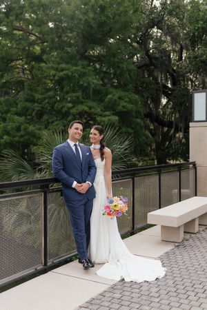 Bride and groom pose on outdoor terrace surrounded by lush greenery, bride holds vibrant bouquet.
