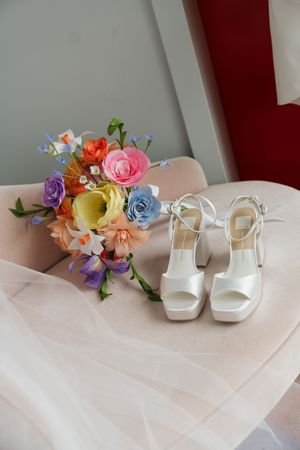 White bridal heels and colorful wedding bouquet with ranunculus and wildflowers on a blush chair.