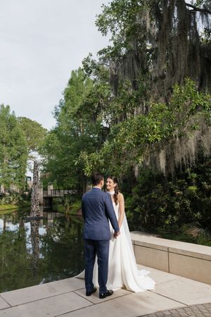 Bride and groom hold hands by a reflective pond surrounded by lush trees draped with Spanish moss.