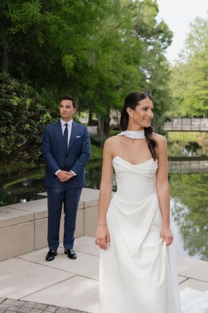 Bride in strapless white gown with halter necklace smiling as groom stands by a reflective pond.
