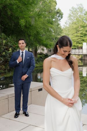 Groom in navy suit gives thumbs up behind bride in white halter gown during outdoor first look by a reflective pond.