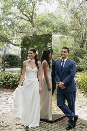 Bride and groom posing outdoors near mirrored sculpture surrounded by lush green trees.