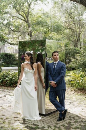 Bride in white gown and groom in navy suit pose beside a mirrored panel in a lush garden setting.