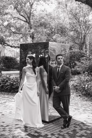 Black and white photo of bride and groom posing beside a mirrored sculpture in an outdoor garden.
