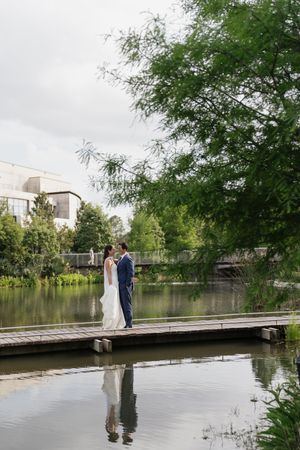 Bride and groom share a romantic moment on a wooden dock, reflected in calm water surrounded by lush greenery.