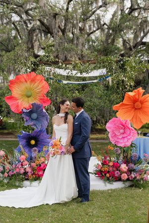 Bride and groom share romantic kiss under vibrant oversized floral wedding arch outdoors.