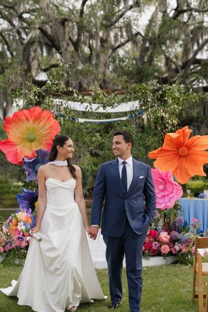 Bride and groom hold hands smiling before colorful oversized paper flowers at outdoor wedding ceremony.