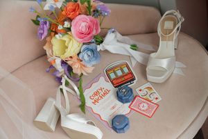 Bridal flat lay with colorful floral bouquet, white heels, wedding invitation, ring boxes, and playing cards on pink chair.