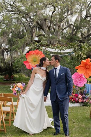 Bride and groom share a kiss at outdoor ceremony with colorful oversized floral decorations and Spanish moss trees.
