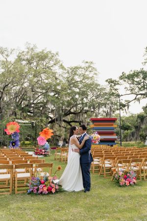 Bride and groom share a kiss at colorful outdoor wedding ceremony with giant paper flowers and rainbow art installation.