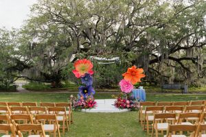 Outdoor wedding ceremony setup with giant paper flowers, wooden chairs, and moss-covered oak tree backdrop.