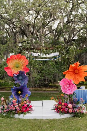 Vibrant oversized paper flower wedding altar with lush floral arrangements under a moss-draped oak tree.