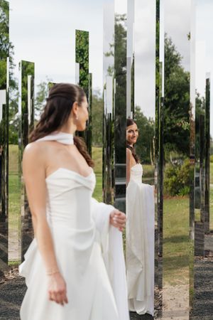 Bride in white halter gown admiring her reflection in tall mirrored columns outdoors in a park.