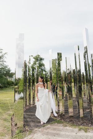 Bride in white strapless gown walks among tall mirrored columns in an outdoor art installation.