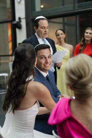 Groom smiling during Jewish wedding ceremony while officiant reads from paper with guests in background.