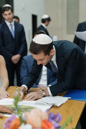 Groom wearing kippah signs Jewish marriage contract at table decorated with colorful flowers.