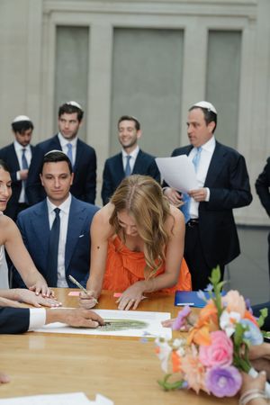 Wedding guests signing ketubah at table with colorful floral arrangement during Jewish ceremony.