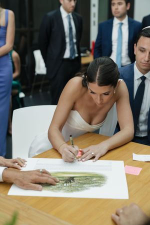Bride in white gown signs ketubah document at wooden table during Jewish wedding ceremony.