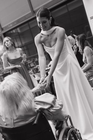 Black and white photo of smiling bride in white gown holding hands with elderly guest in wheelchair.