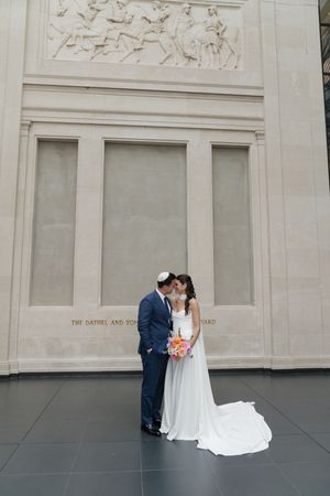 Bride and groom kiss inside a grand museum hall with classical stone relief architecture and long bridal train.