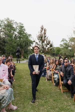 Groom in dark suit walks down outdoor wedding aisle carrying a book while guests seated in wooden chairs look on.