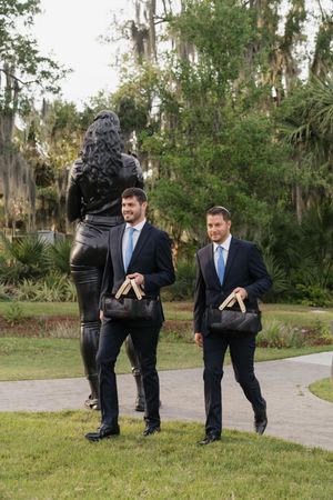 Two groomsmen in navy suits with blue ties carry black bags while walking with a woman in a park.