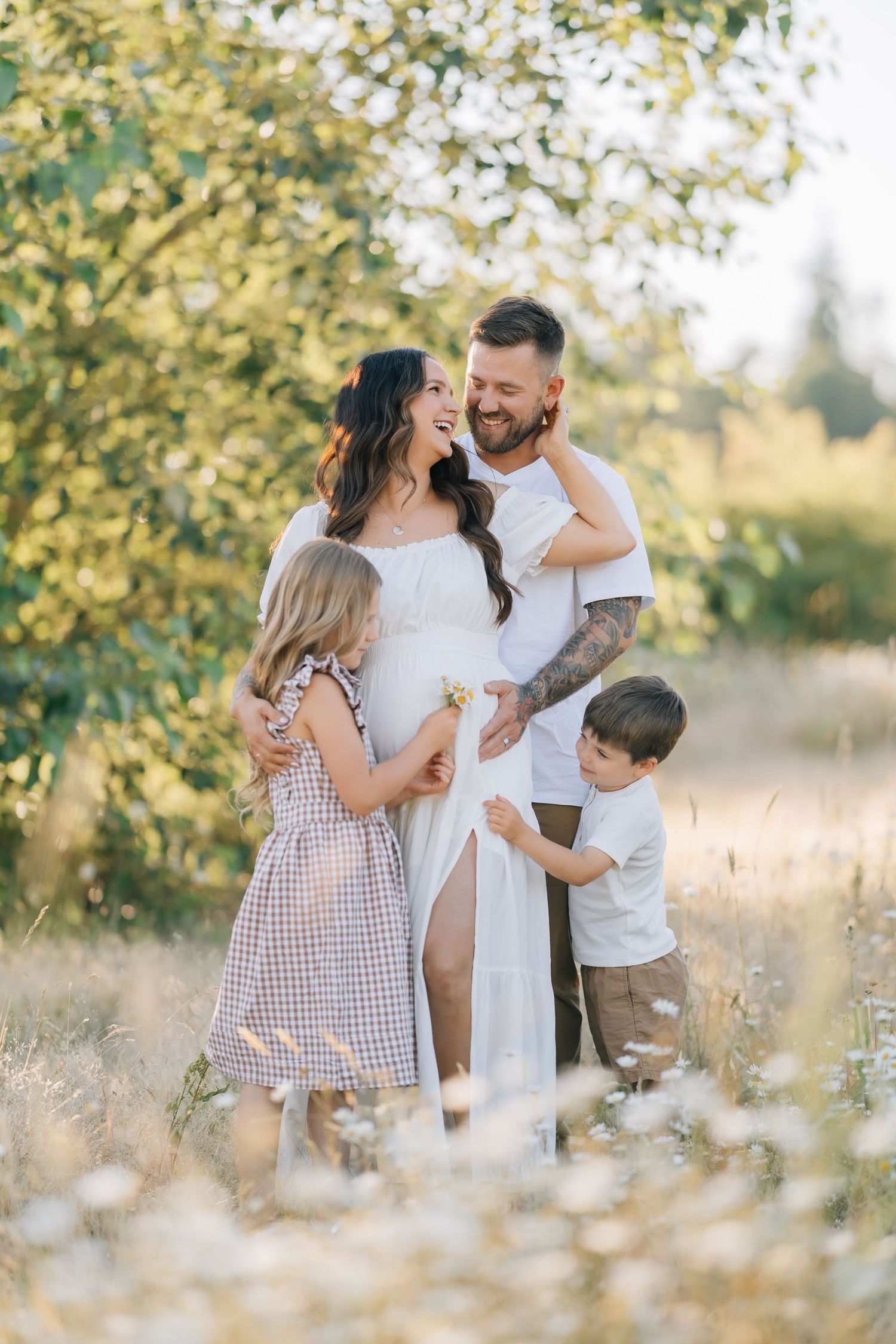 Happy pregnant family with two children posing in a sunny wildflower meadow, mom in white dress, dad with tattoos smiling.