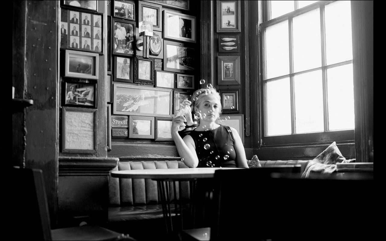Black and white photo of a woman sitting alone at a wooden desk in a vintage room with framed pictures on the wall.