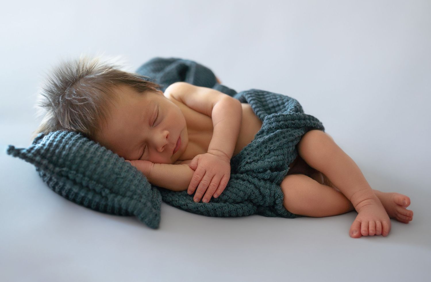 Newborn baby sleeping peacefully on a teal knit blanket and pillow in a serene studio photo.