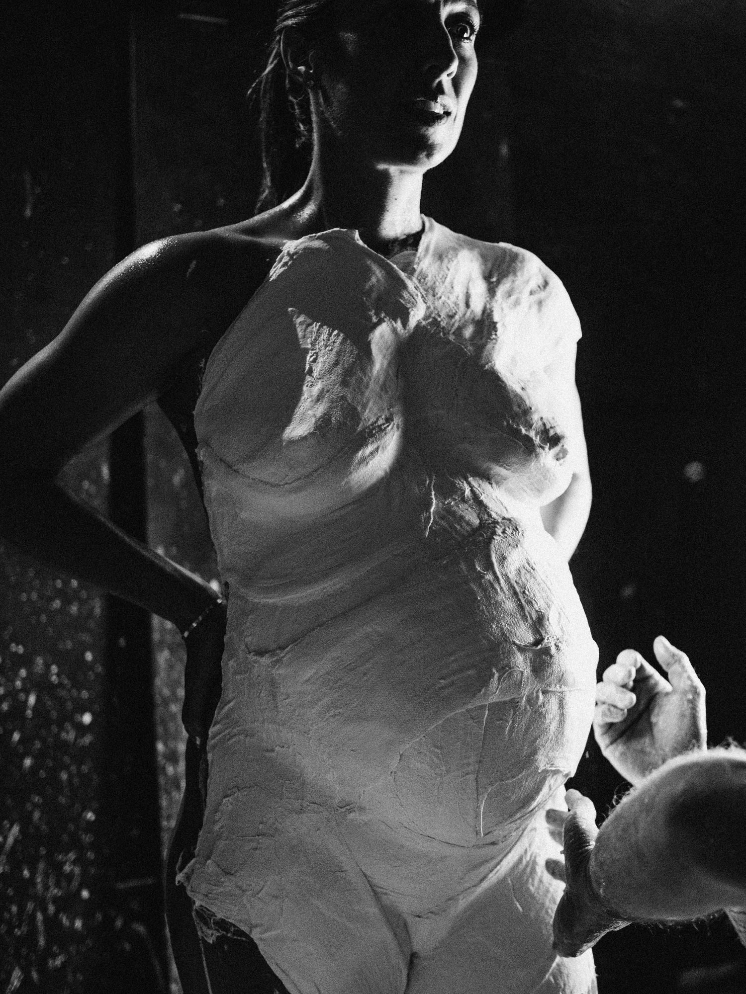 Black and white photo of a pregnant woman covered in white plaster, posing dramatically in dark studio lighting.